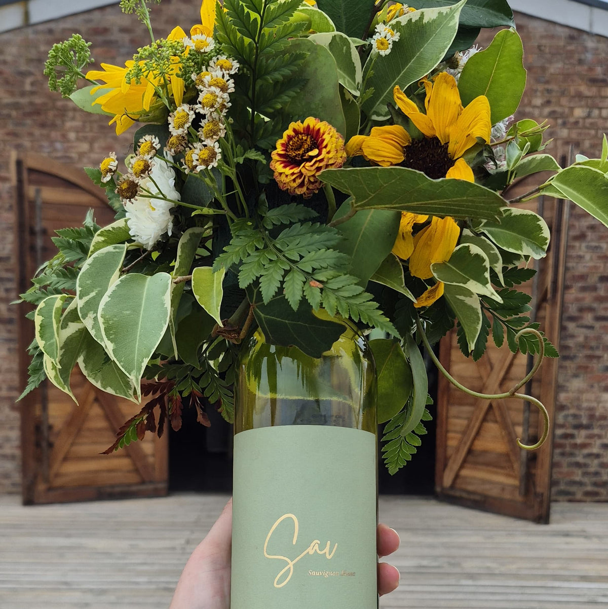 Bouquet of flowers in a green bottle held by a person against a brick wall.